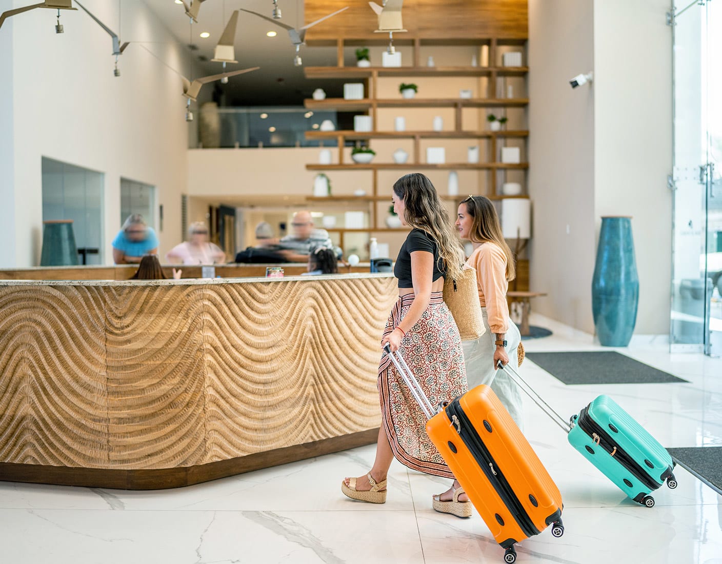 Two women carrying luggage walk through a stylish hotel lobby, surrounded by modern decor and soft lighting.
